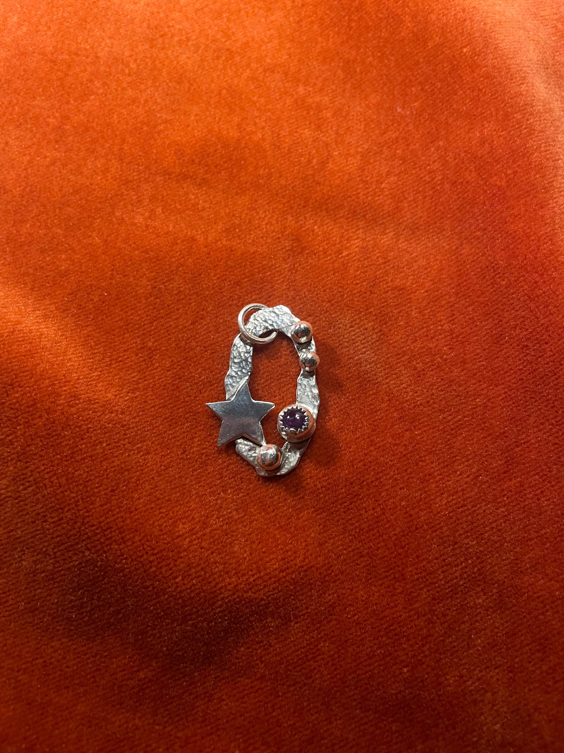 Silver charm with star and circular celestial design and amethyst stone set inside on an orange velvet background.