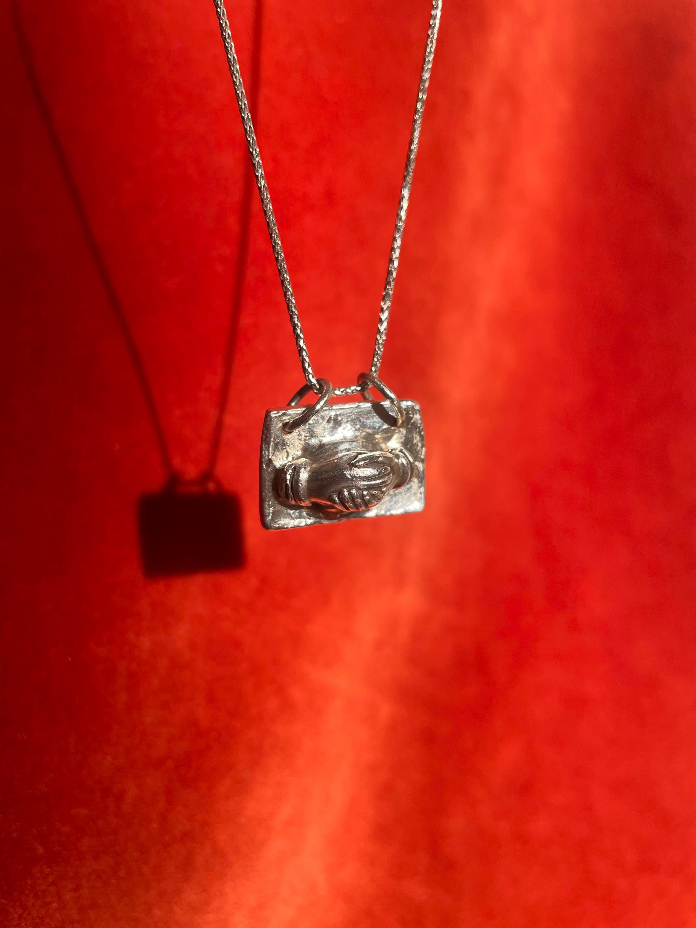 Silver necklace with a pendant of hands holding each other on a red background.