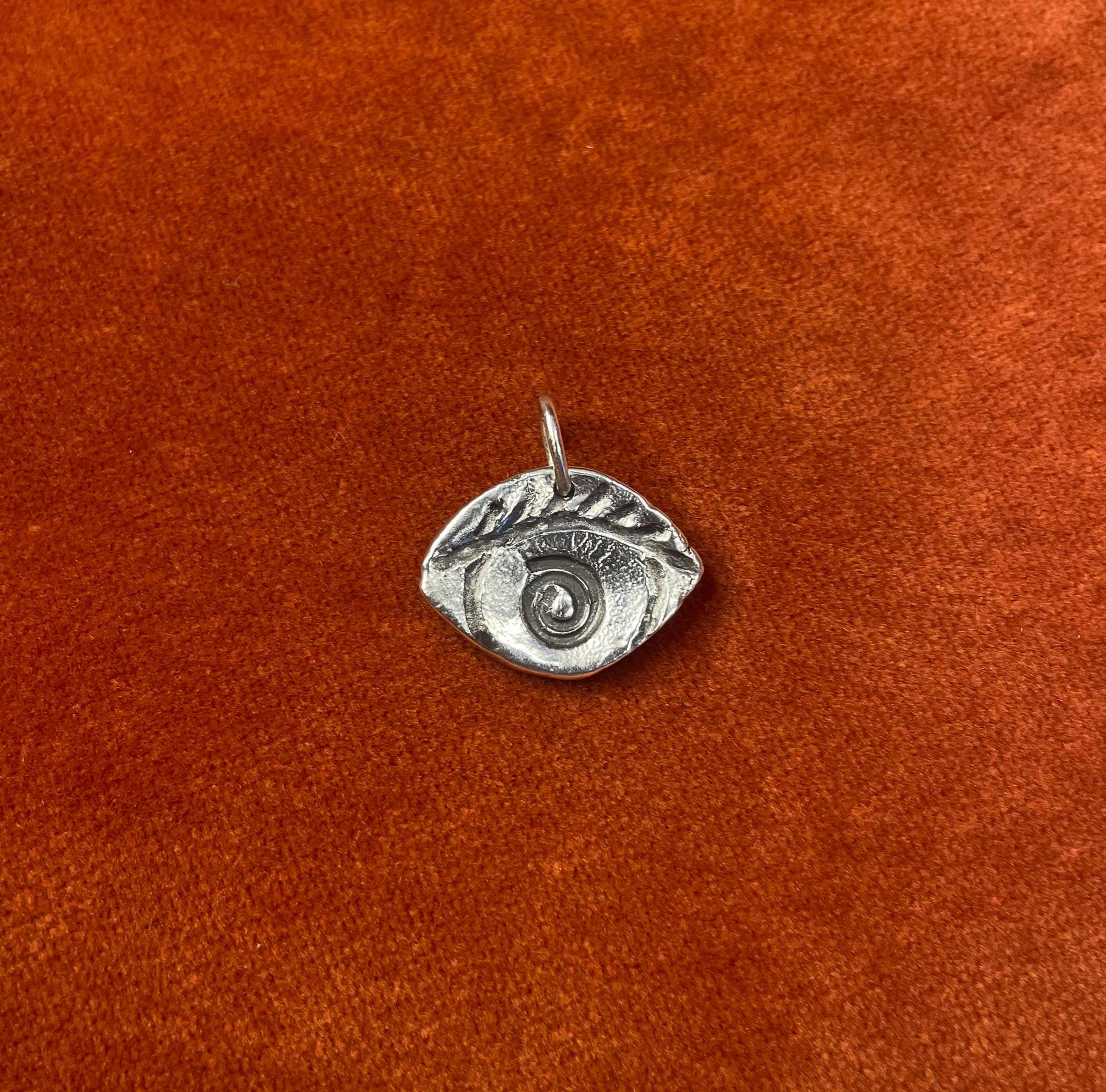 Silver eye-shaped charm laying on an orange velvet fabric background.