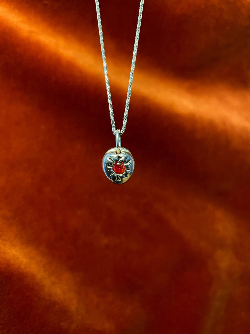 Silver necklace with a round pendant featuring a red gemstone on an orange fabric background