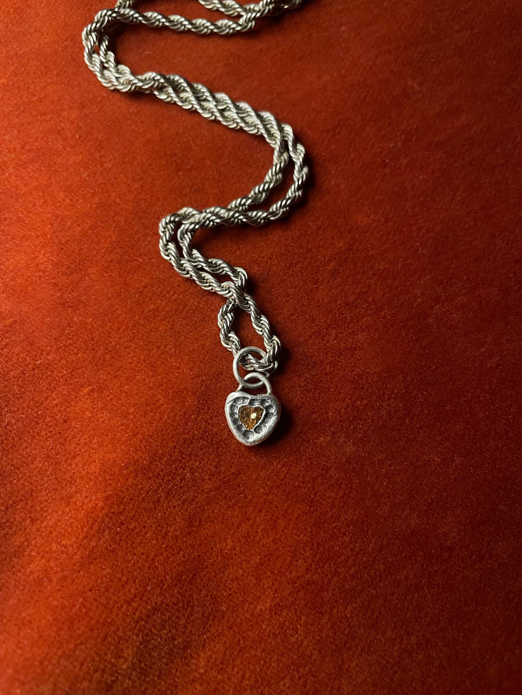 Silver roped chain with a heart-shaped pendant on an orange velvet background.