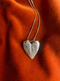 Silver heart-shaped pendant on a chain against an orange fabric background.