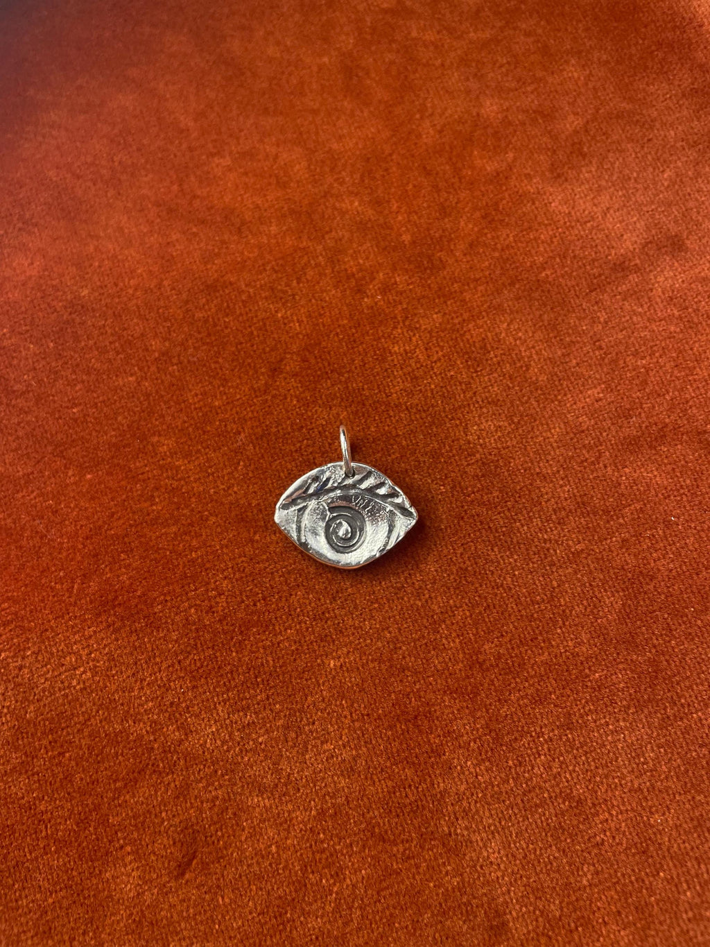 Silver eye-shaped charm with a spiral in the center laying on a orange fabric surface.