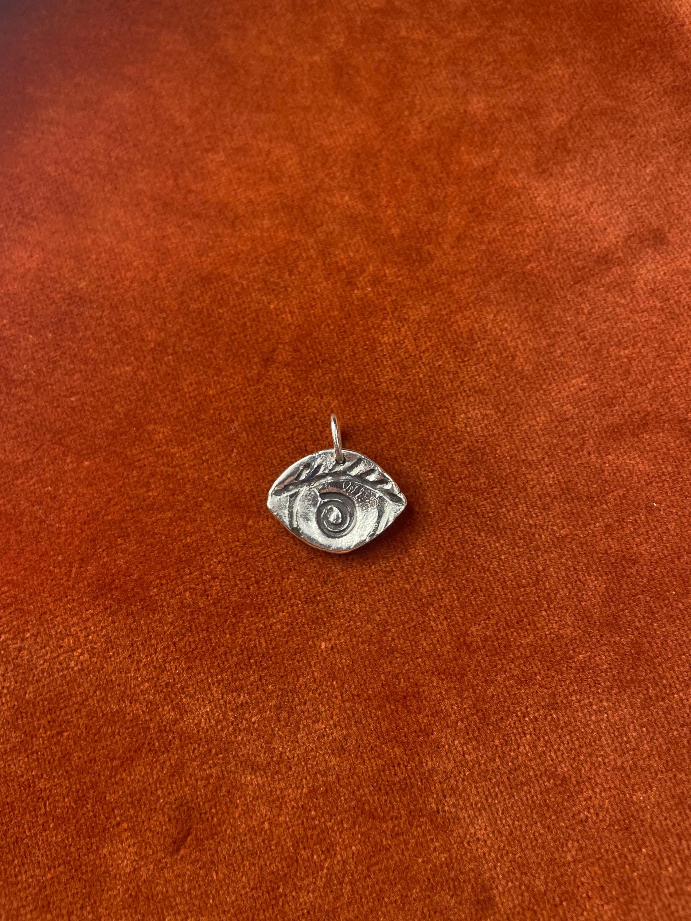 Silver eye-shaped charm with a spiral in the center laying on a orange fabric surface.
