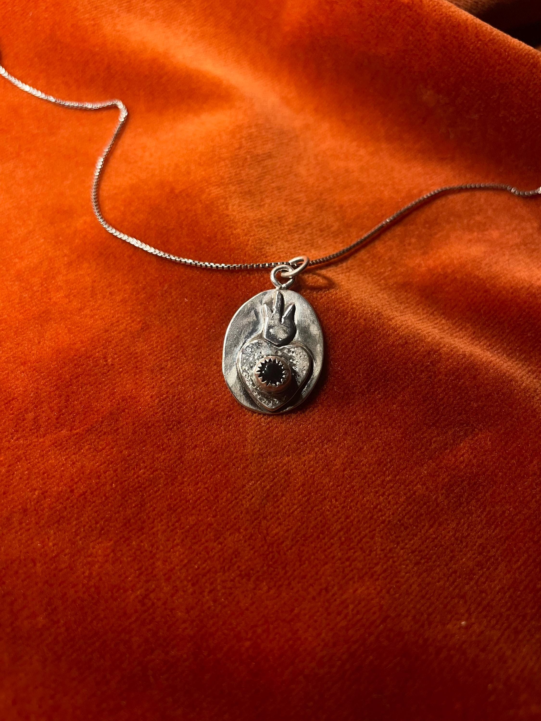 Silver chain with a sacred heart charm and an amethyst stone set in the center. Staged on an orange velvet background.