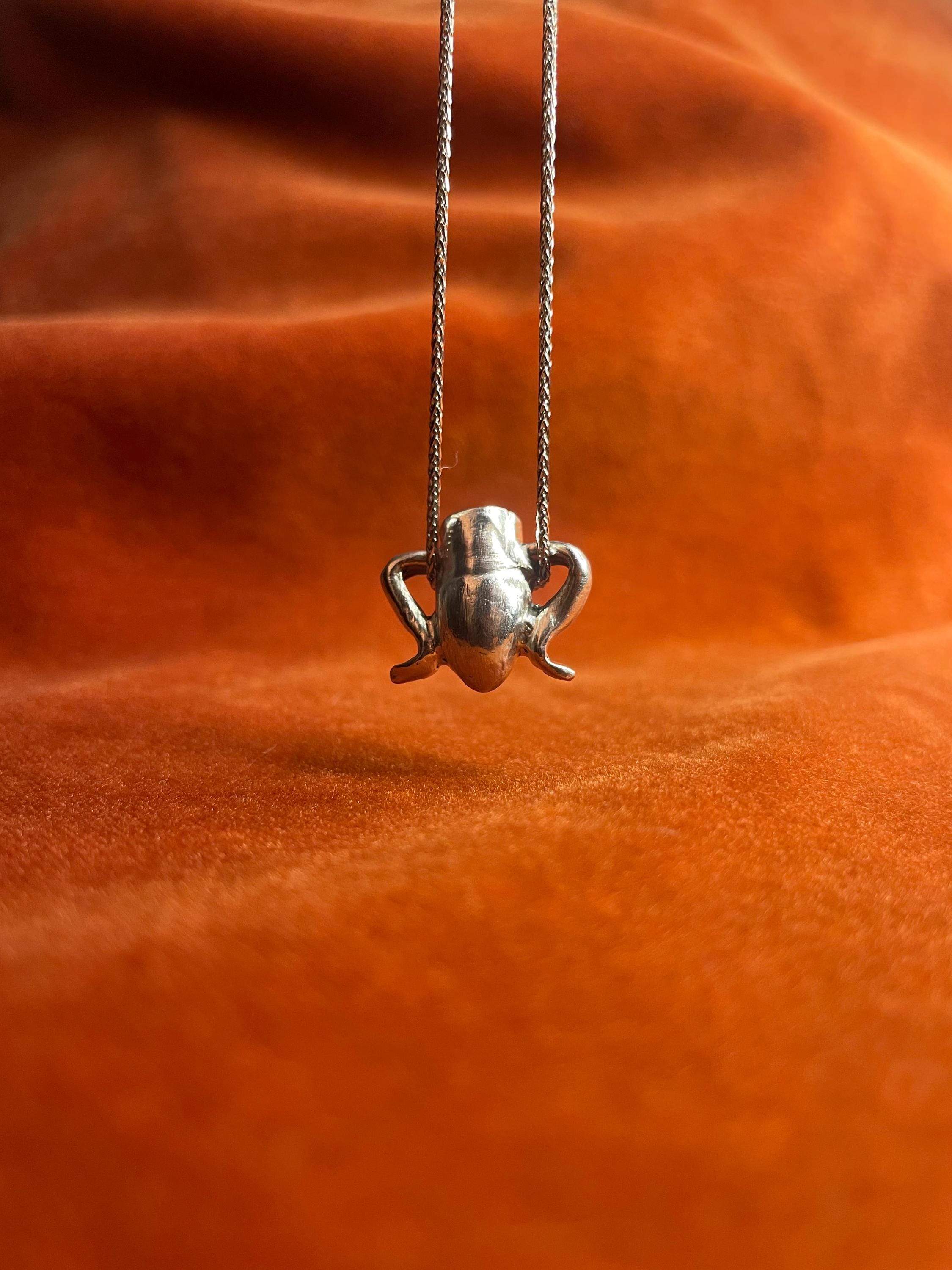 Silver chain with a small vase charm dangling above an orange fabric background.