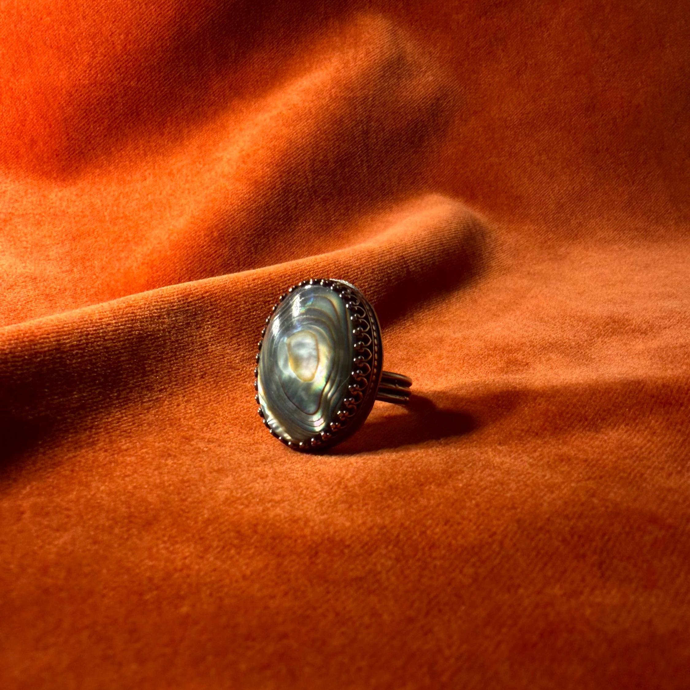 Silver ring with a colorful abalone oval shaped stone set in an ornate bezel sitting on a medium sized band. Staged on an orange velvet background.