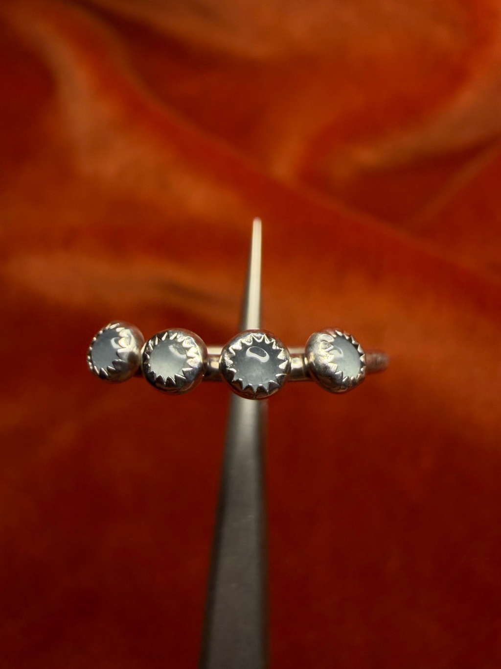 Close up image of silver ring with four small milky blue aquamarine stones, set in serrated bezels. Laid on a small thing band. Ring is held by small tweezers.