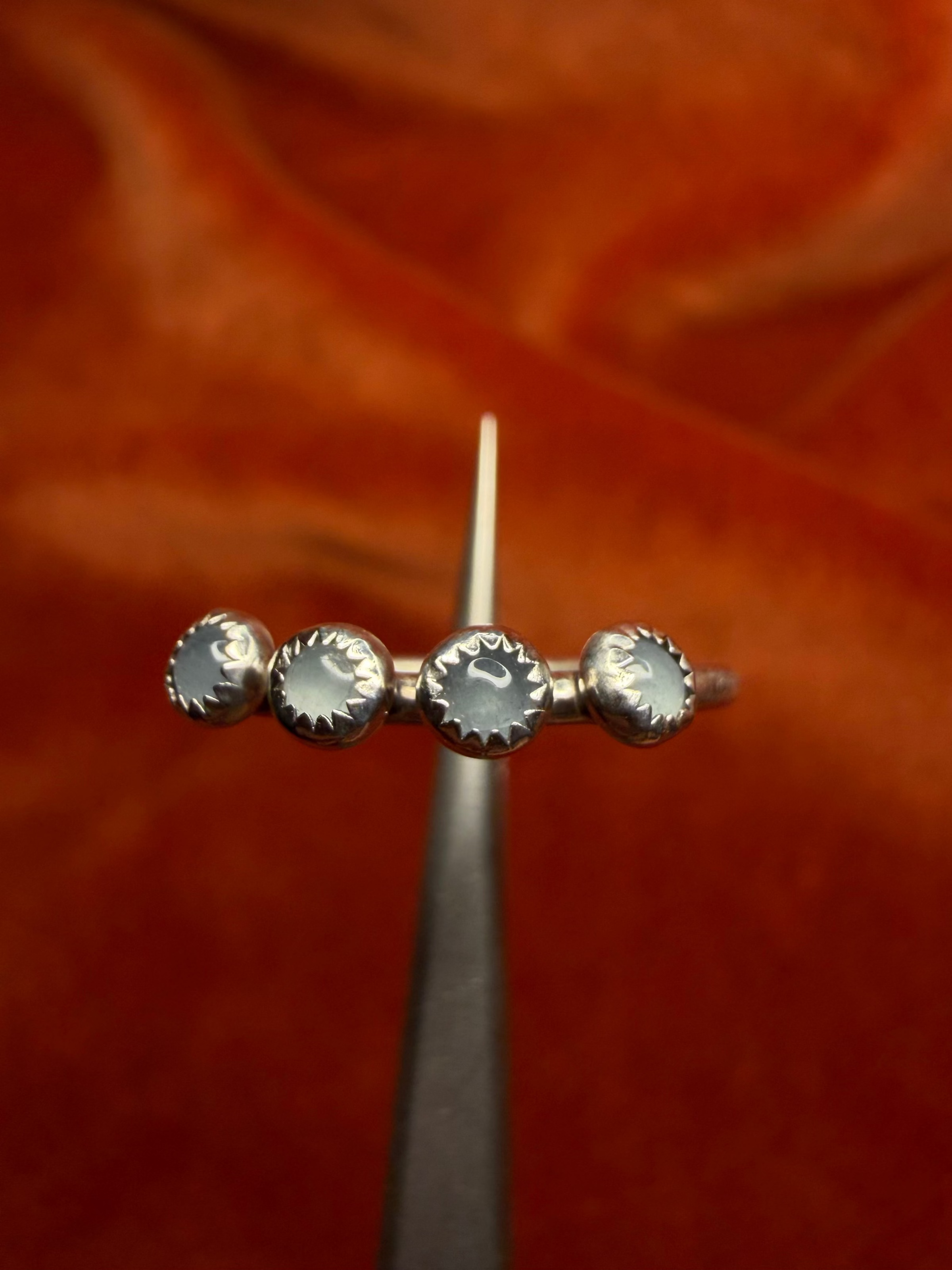 Close up image of silver ring with four small milky blue aquamarine stones, set in serrated bezels. Laid on a small thing band. Ring is held by small tweezers.
