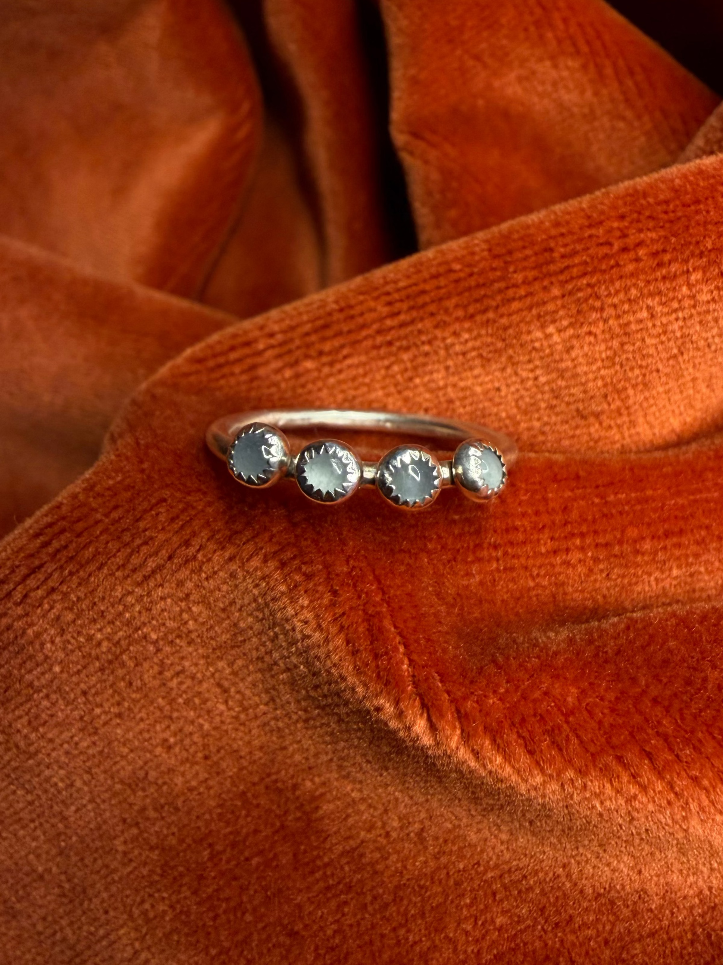 Silver ring with four small milky blue aquamarine stones, set in serrated bezels. Laid on a small thing band. Ring is staged on an orange velvet background.