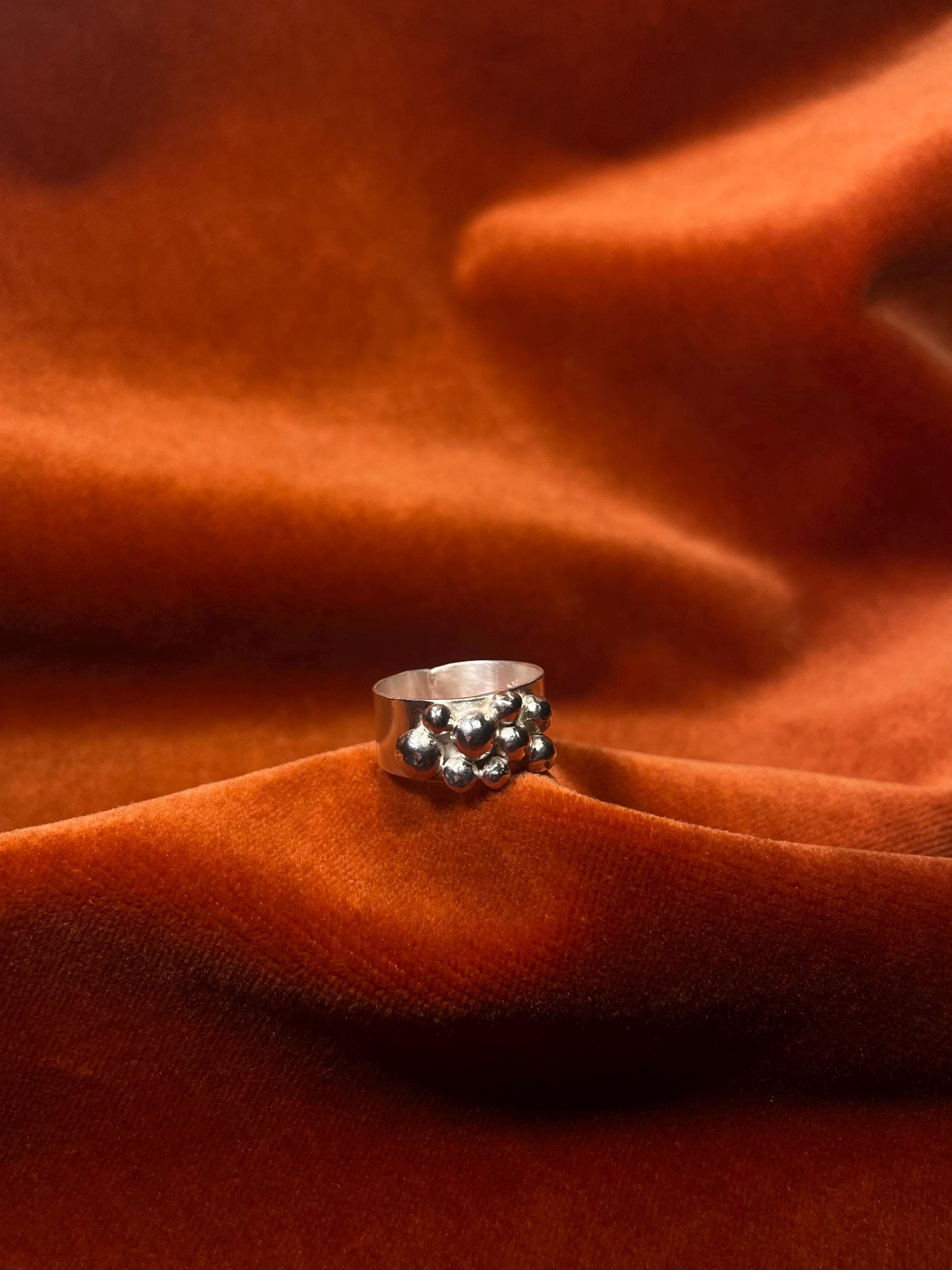 Silver ring with small bubbles forming on the top of the ring. Laying on a velvet orange backdrop.