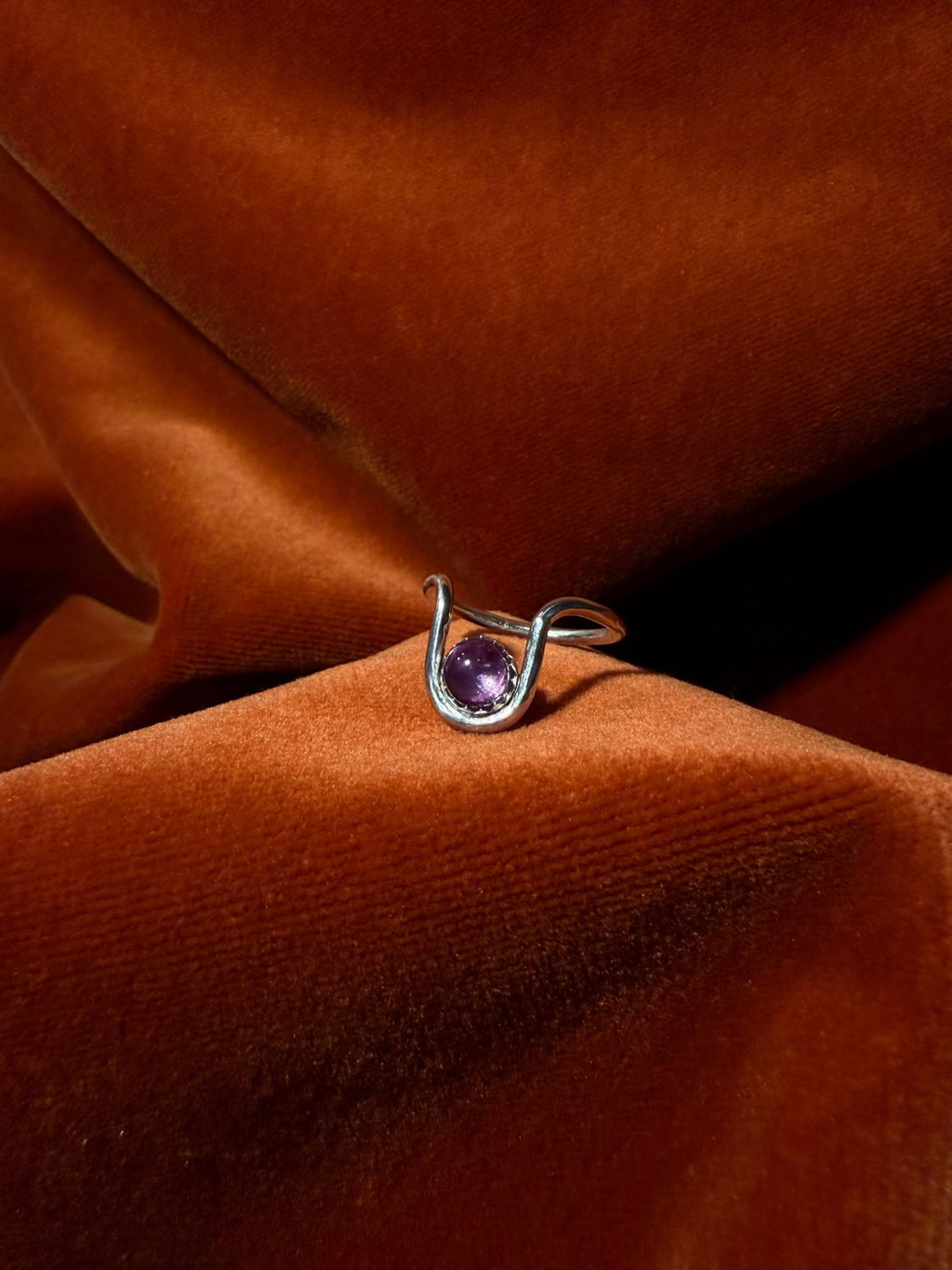 Close up image of a silver ring with bend in the ring where a serrated bezel holds a pink sapphire stone. Staged against an orange velvet background.