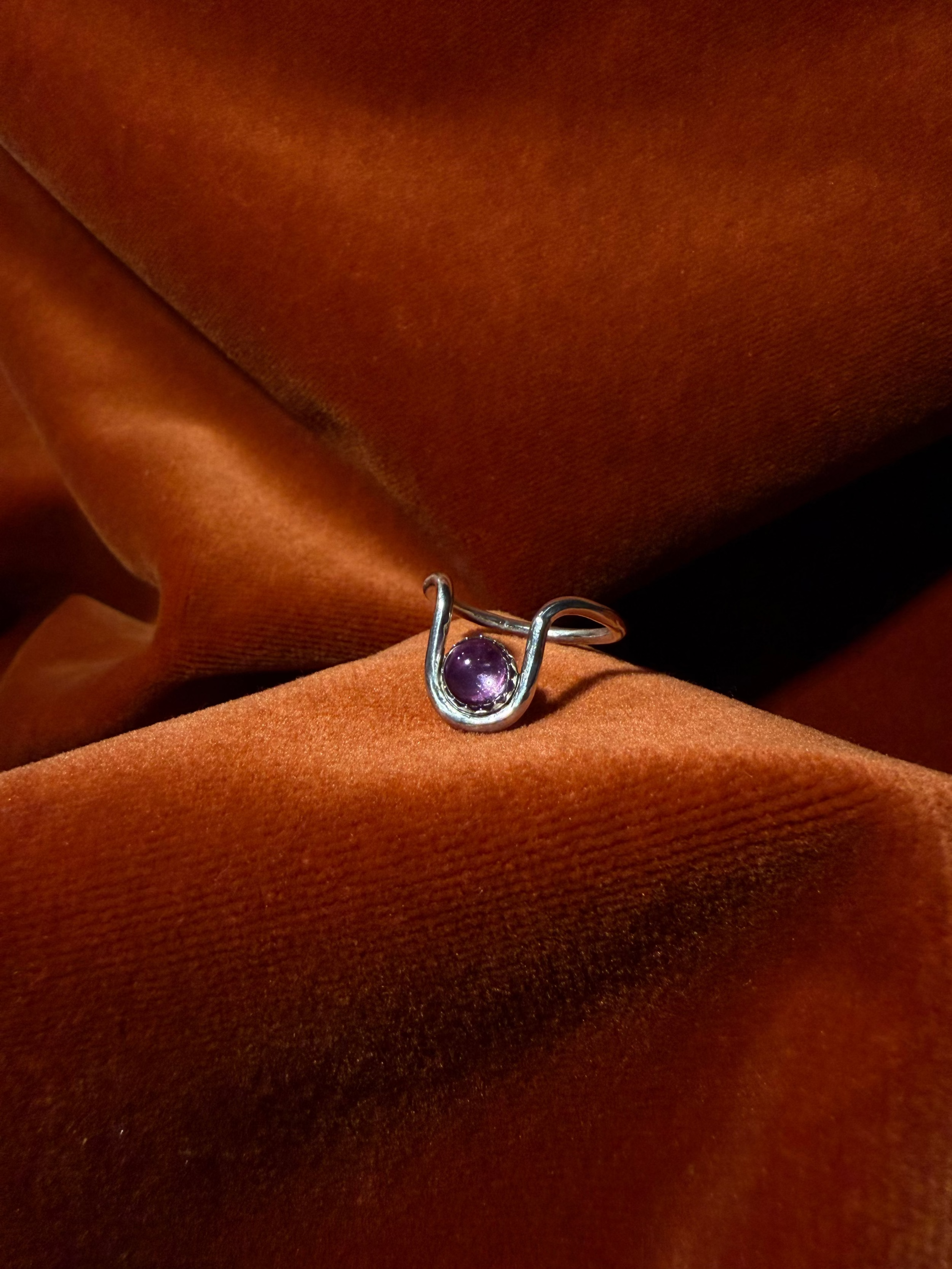 Close up image of a silver ring with bend in the ring where a serrated bezel holds a pink sapphire stone. Staged against an orange velvet background.