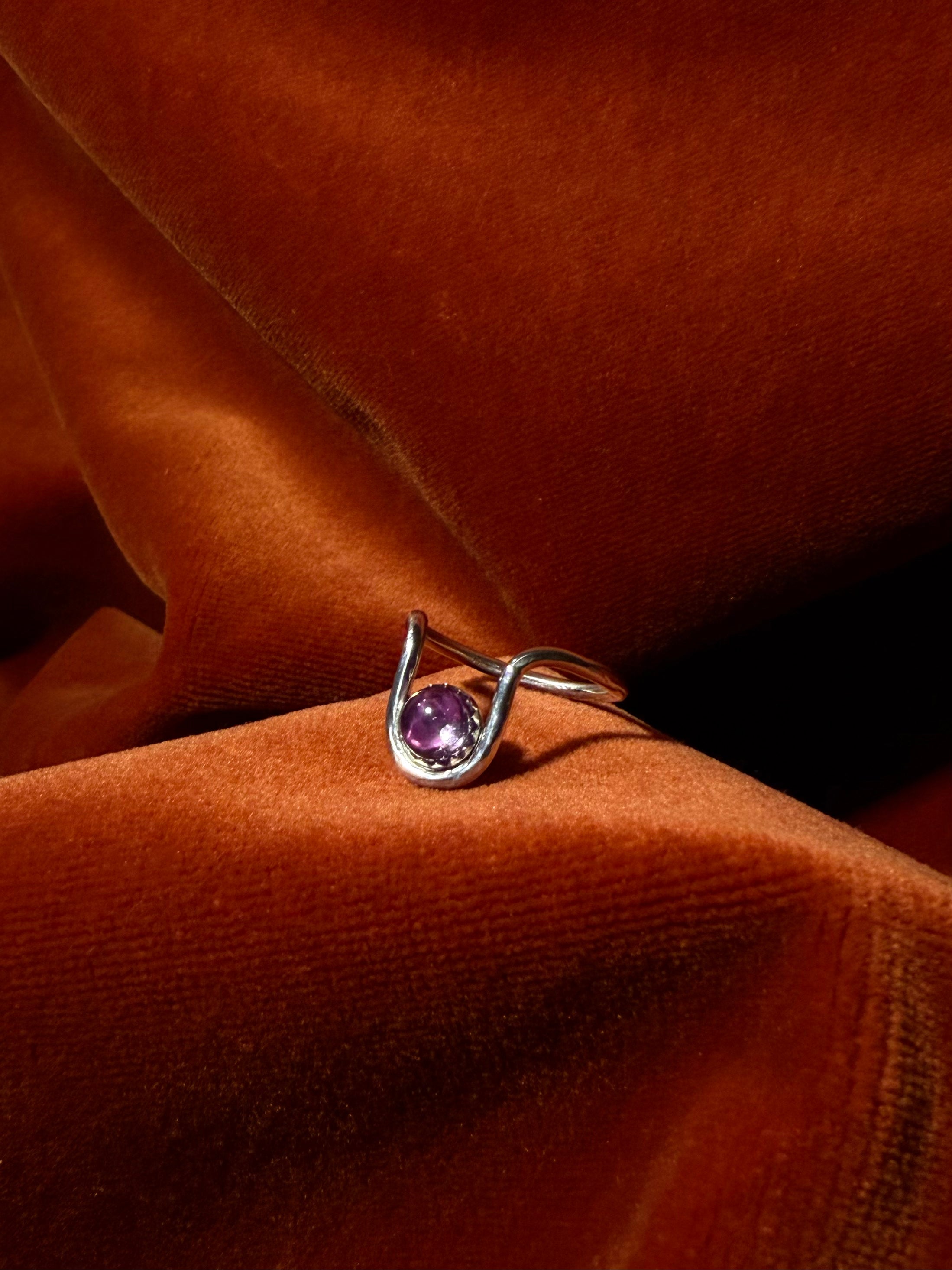 Close up image of a silver ring with bend in the ring where a serrated bezel holds a pink sapphire stone. Staged against an orange velvet background.