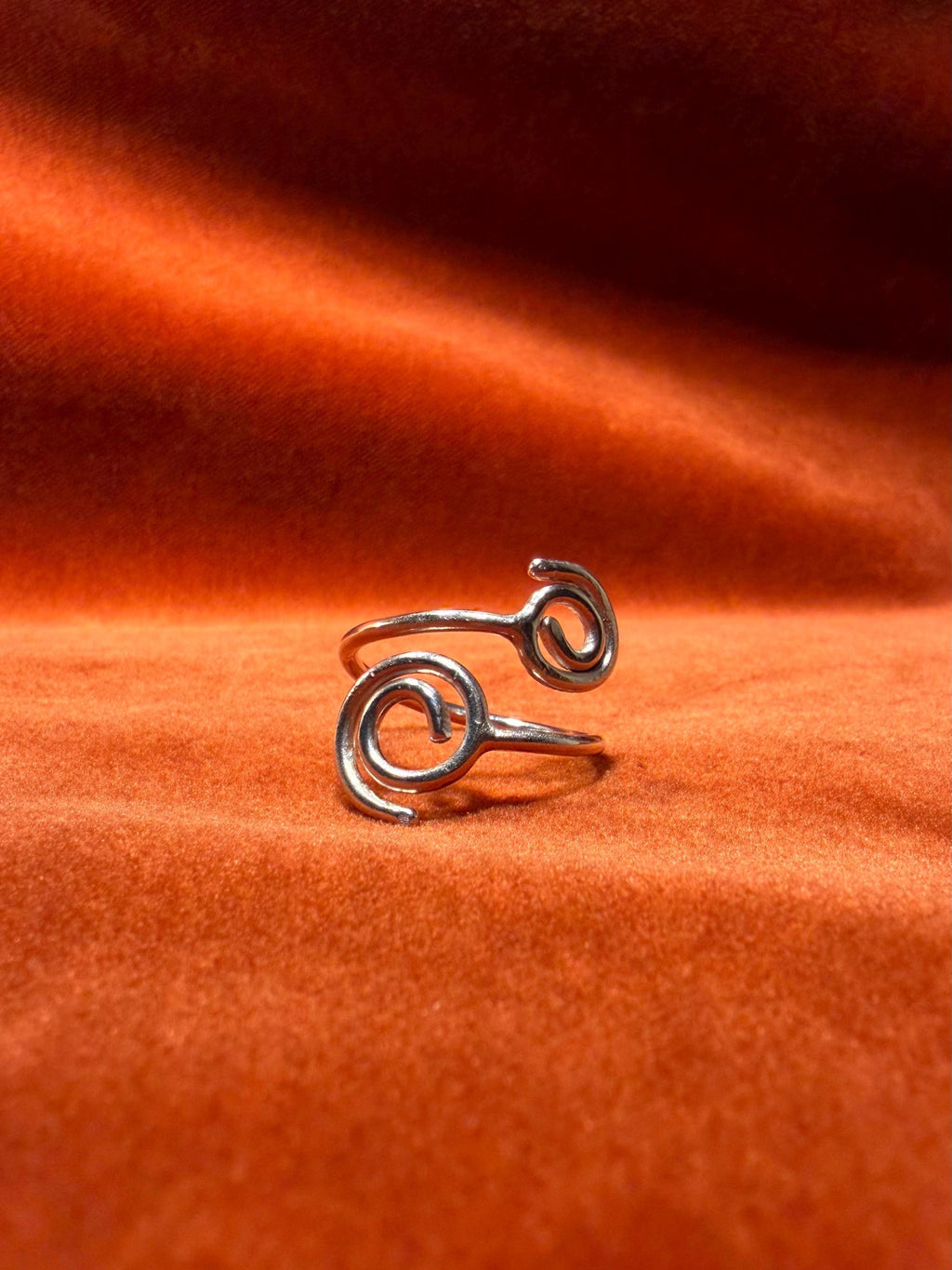 Silver ring with spirals on each end of the ring. The is staged against an orange velvet backdrop. 