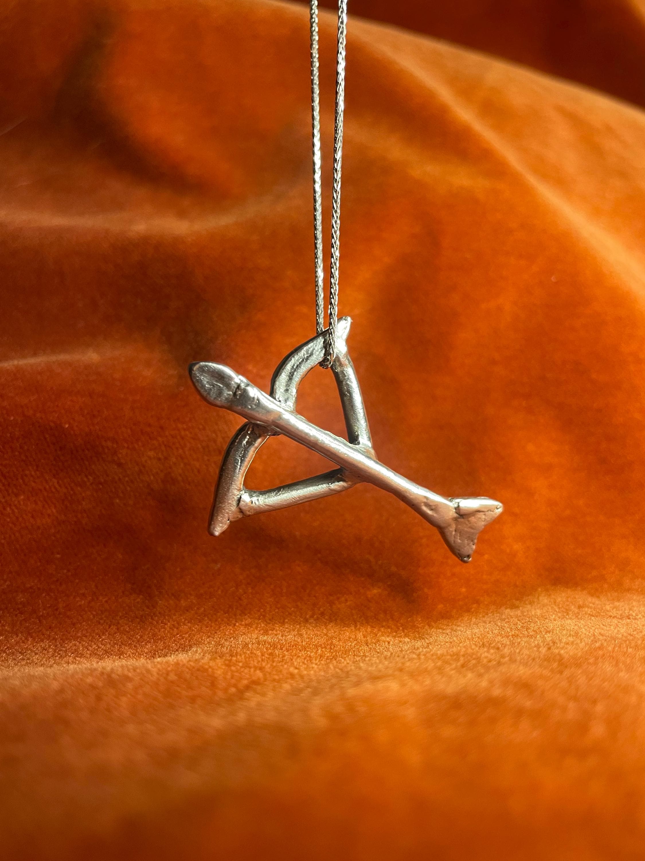 Silver bow and arrow charm dangling on a silver chain. Staged against an orange velvet background.