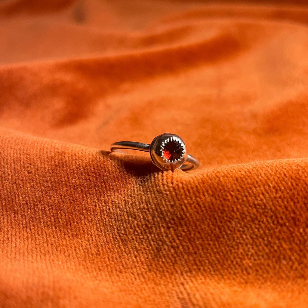 Silver ring with a serrated bezel enclosure and a rose cut garnet stone set inside sitting on a thin band. Ring is staged on an orange velvet background.