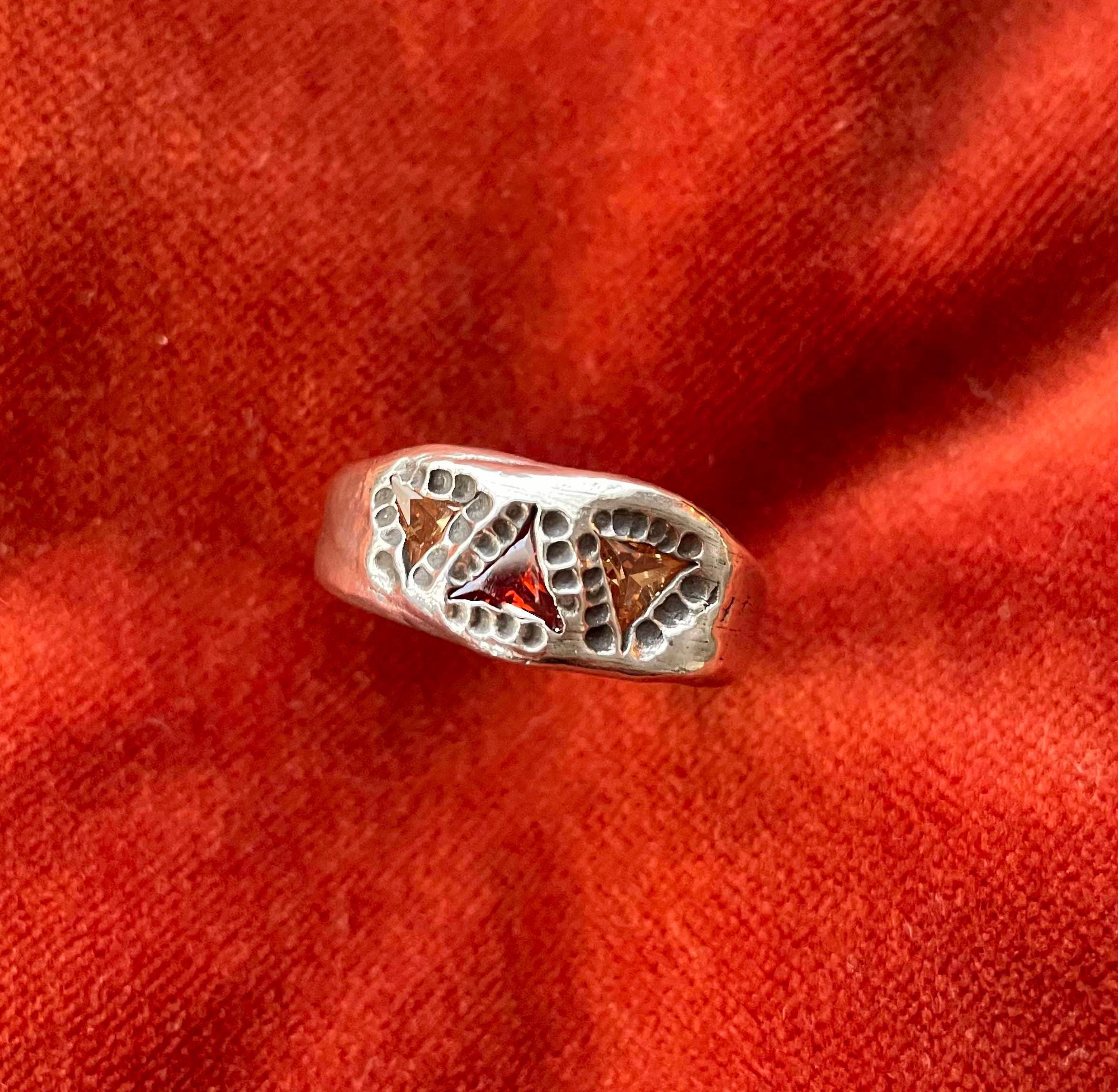 Top down view of a silver ring with three cubic zirconia triangle shaped stones. Two stones are yellow and surround one red stone. Oxidized circular impression surround the stones. 