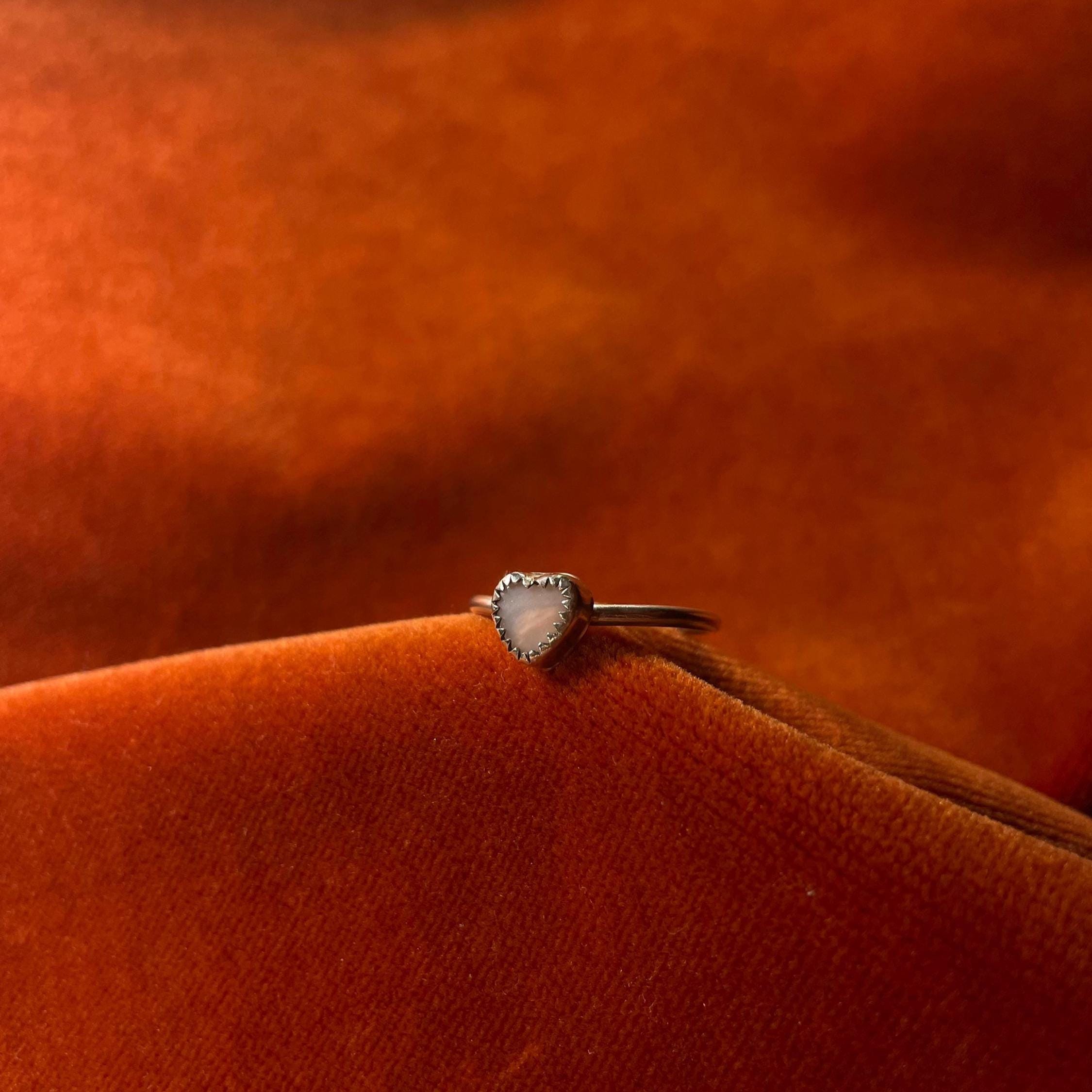 Silver ring on an orange velvet background. A pink opal heart shaped stone set in a serrated bezel sits on a thin band.