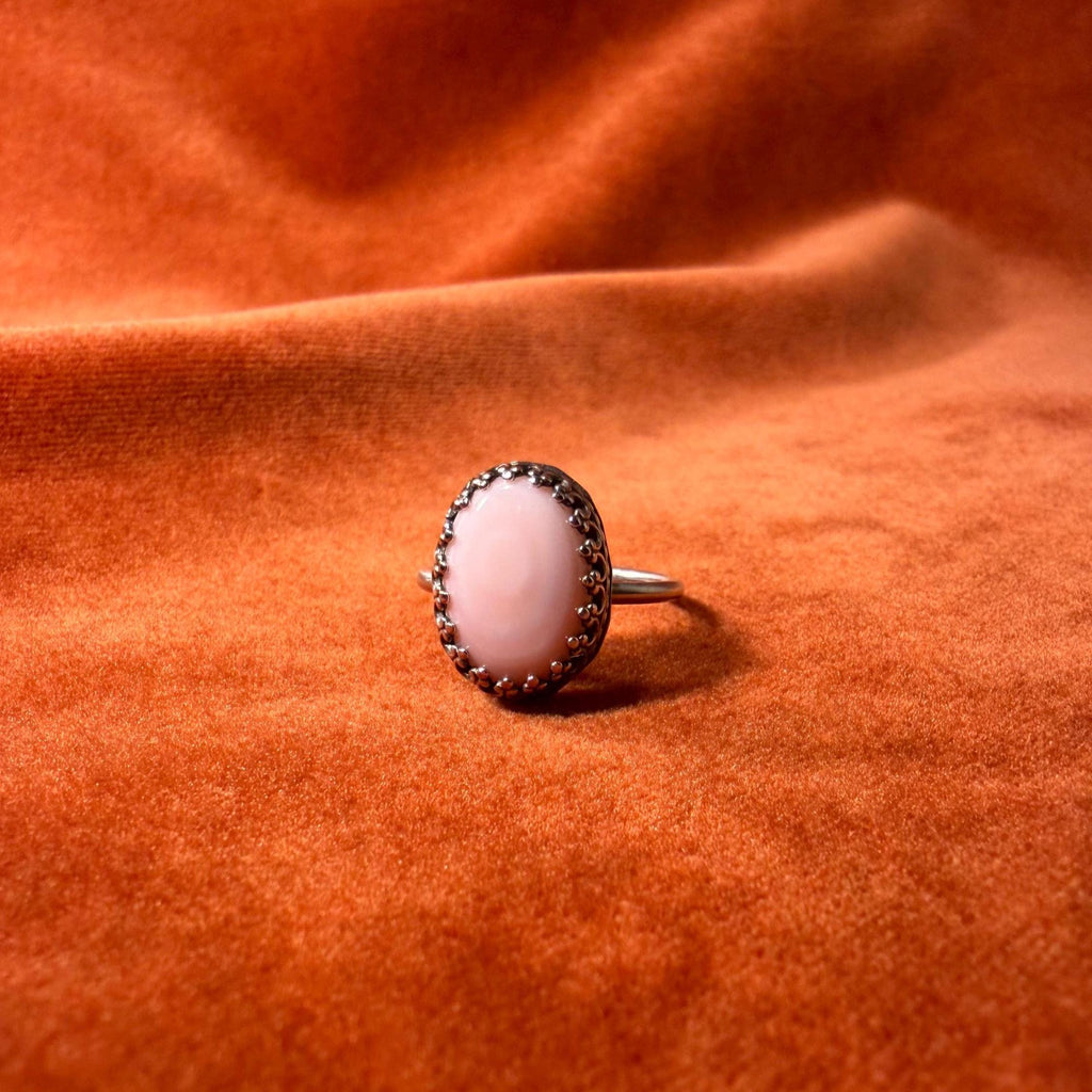 Silver ring with a pink conch stone set in an ornate bezel on a thin band. Set against an orange velvet background.