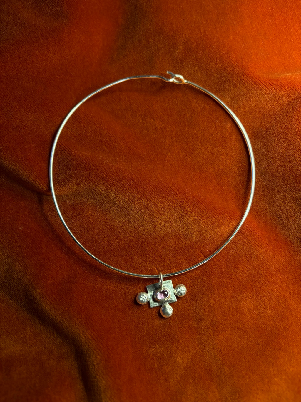 Silver charm on a silver wire choker necklace. Charm features a pink sapphire stone set in the center with three silver orbs. Stage on a orange velvet background.