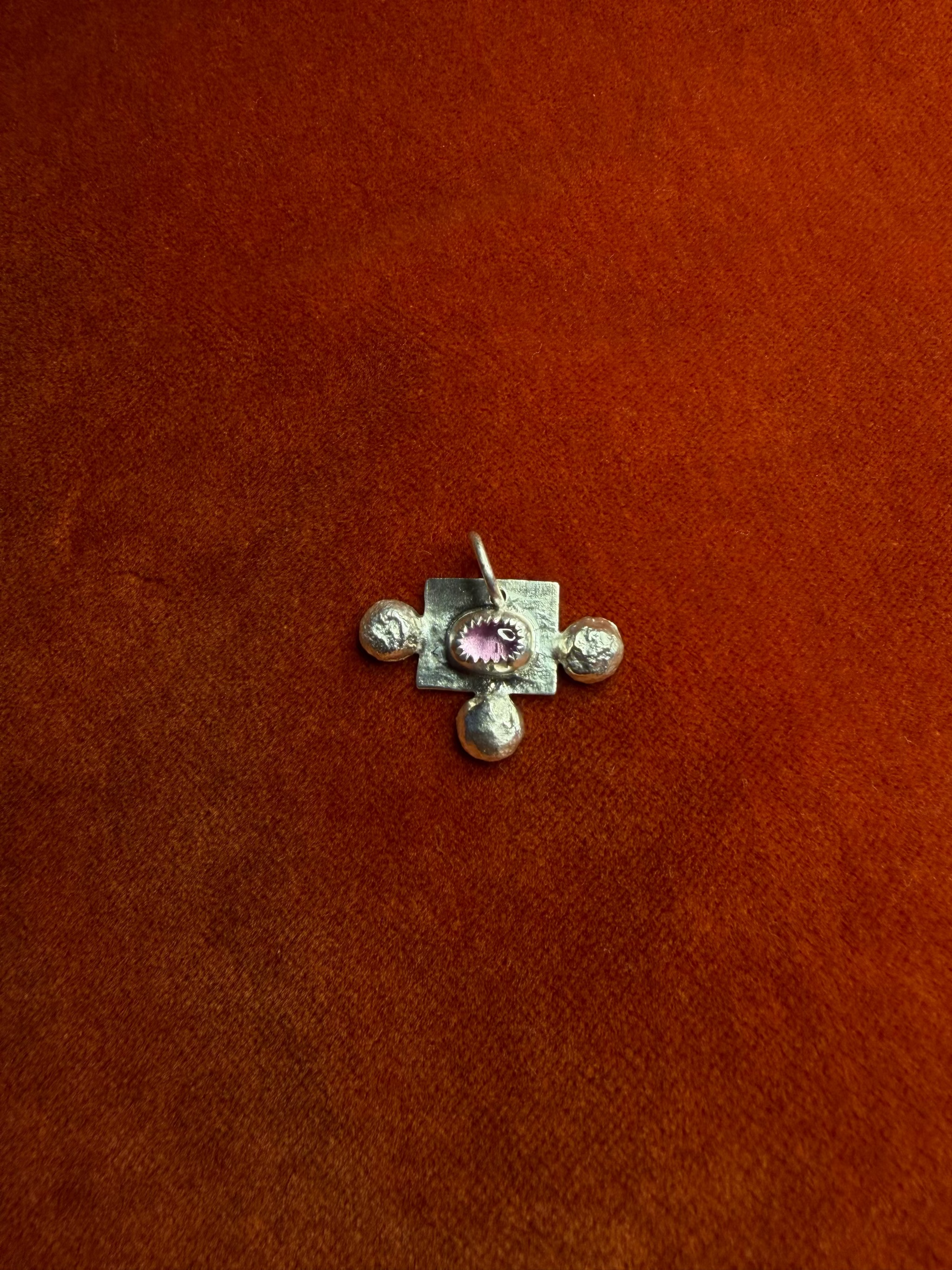 Image of a silver charm featuring a pink sapphire stone set in the center with three silver orbs. Stage on a orange velvet background.