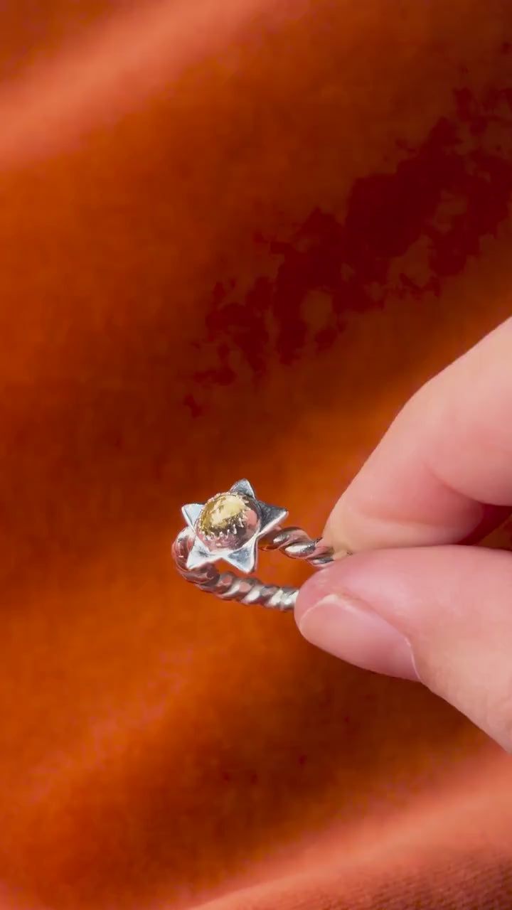Video of a models hand hold a silver ring with a rope band. A star with a golden citrine stone set in the center of the star. Staged on an orange velvet background.