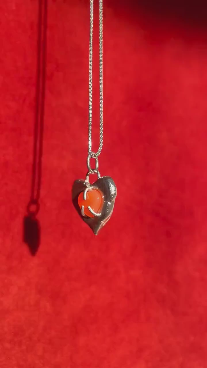 Video of a silver heart charm with a carnelian stone set in the center. Charm dangles on a silver necklace.