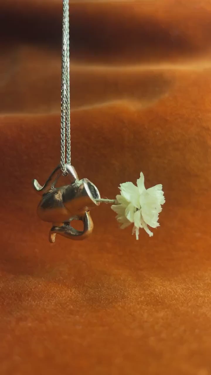 Video of a silver vase dangling on a silver chain. Small flower sits in vase.