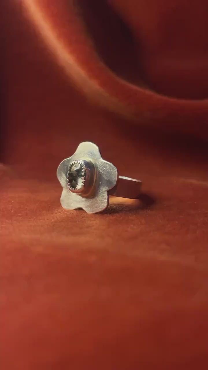 A moving video rotating around a silver ring with a flower petal design and a moss agate gemstone set in the center sitting on a small band. Staged against an orange velvet background.