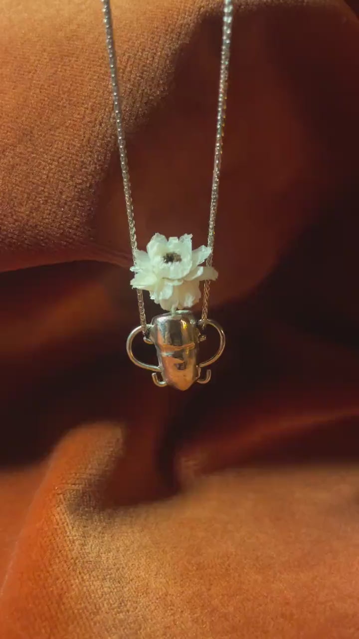 Video of a silver vase pendant dangling from a silver chain. Tiny flower sits inside vase. 