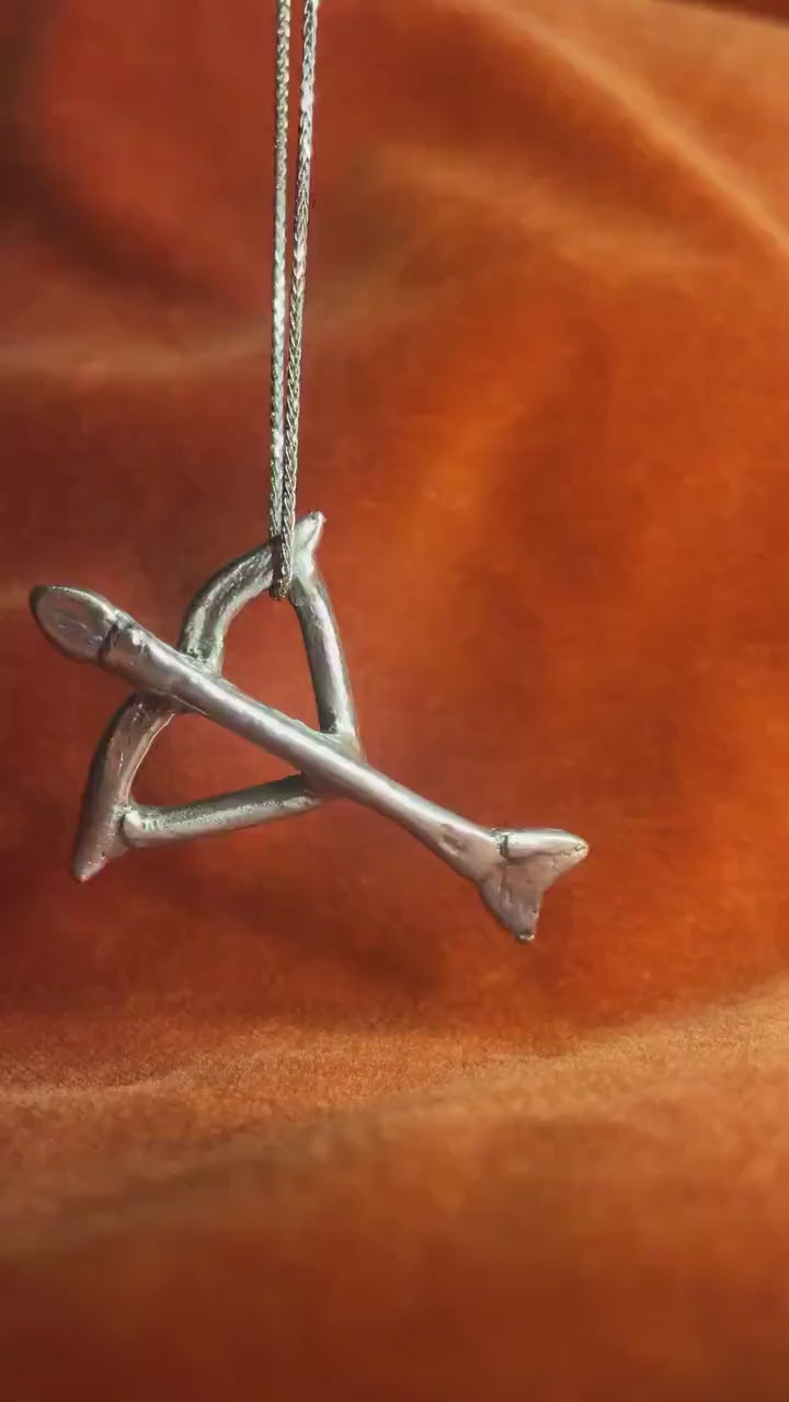 Video of a silver bow and arrow charm swaying left and right on a silver chain against an orange velvet background.