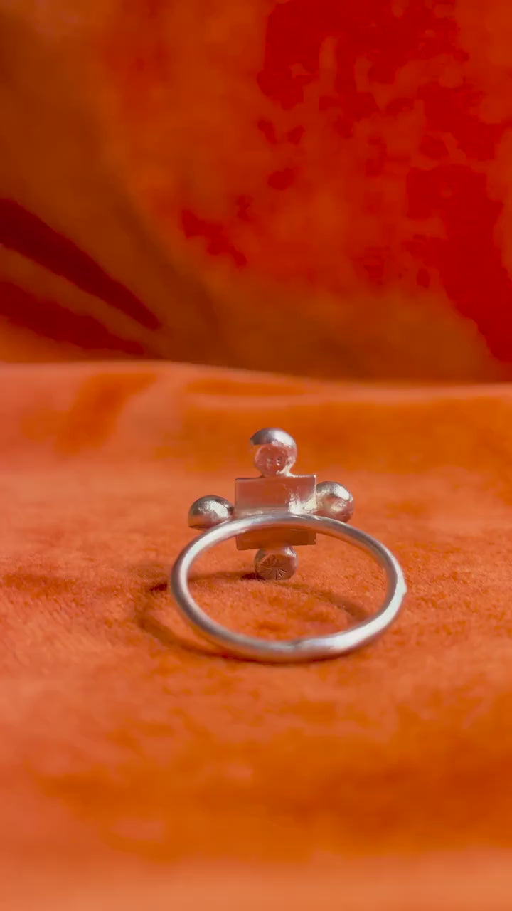 A model's hand picks up a silver ring with four orbs set around a small square shape design sitting on a thin band. Staged against an orange velvet background.
