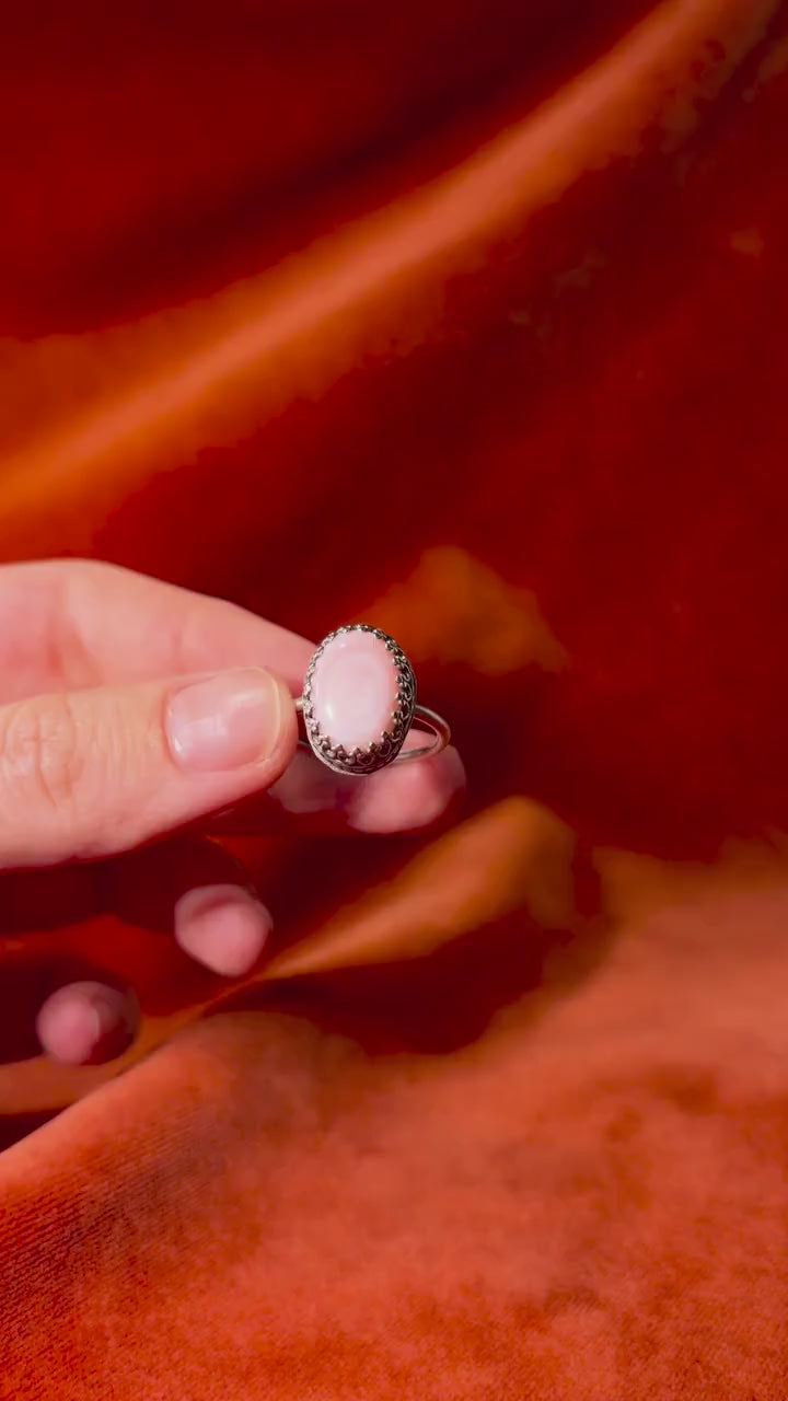 A model's hand holds a silver ring with a pink conch stone set in an ornate bezel on a thin band. Set against an orange velvet background.