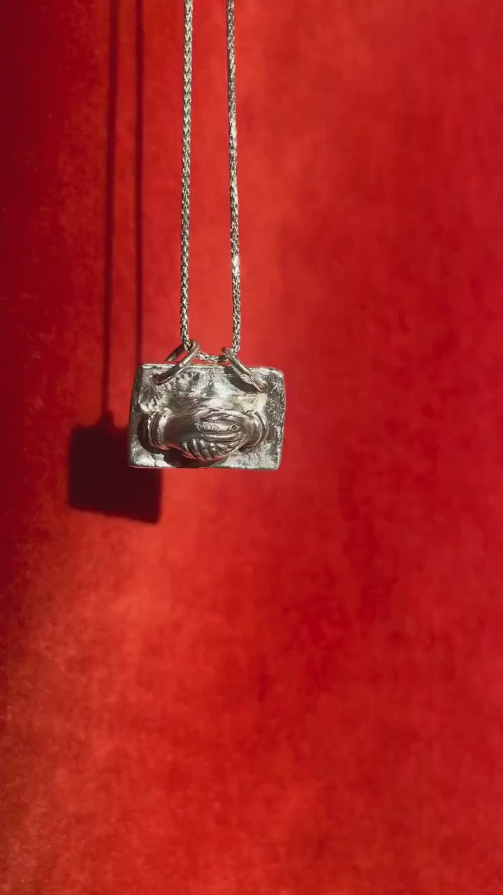 Video of a silver charm with hands holding dangling on a silver necklace.