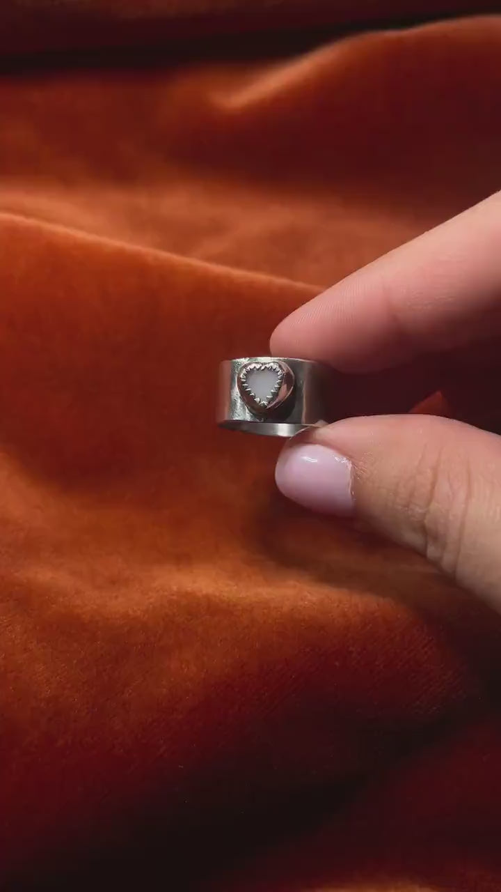 A model's hand holding a silver ring with a pink opal stone set on a chunky band. Staged on an orange velvet backdrop.