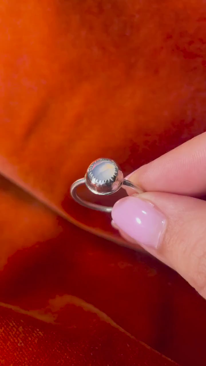 A model's hand holding a silver ring with a sky blue chalcedony rose cut cabochon round stone set in a serrated bezel on a thin band. Set against an orange velvet background.