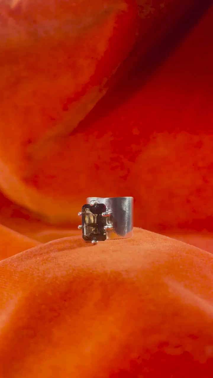 Silver ring with a smoky quartz emerald cut stone, set within five prongs on top of the large band. A models hand comes in and picks up the ring as it shines in the light.
