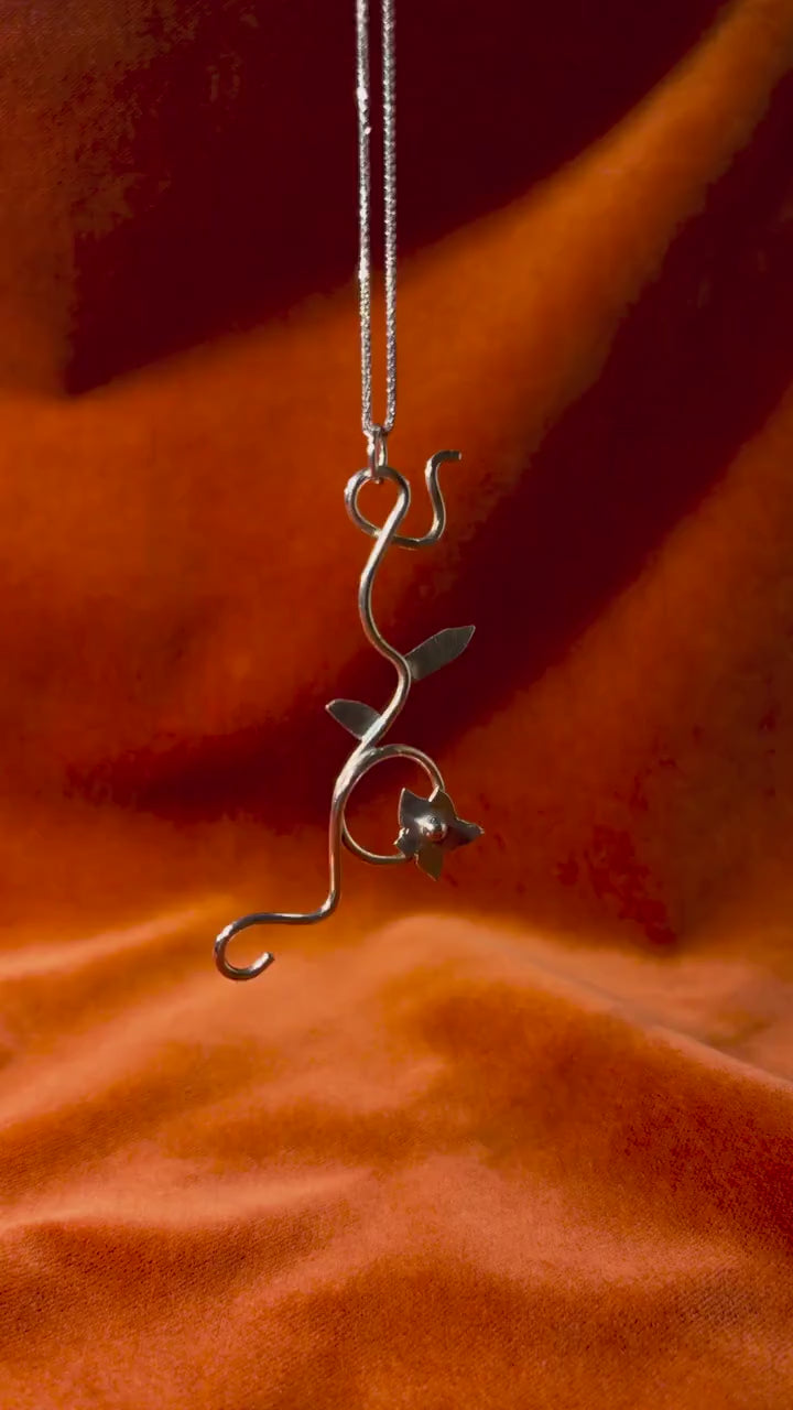 Silver jasmine flower vine charm dangling from a silver chain against an orange velvet background.