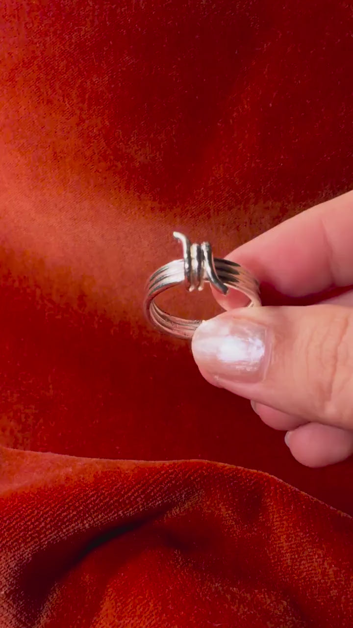 Video of a model's hand holding a silver ring featuring a barbed wire design. Staged on an orange velvet background.
