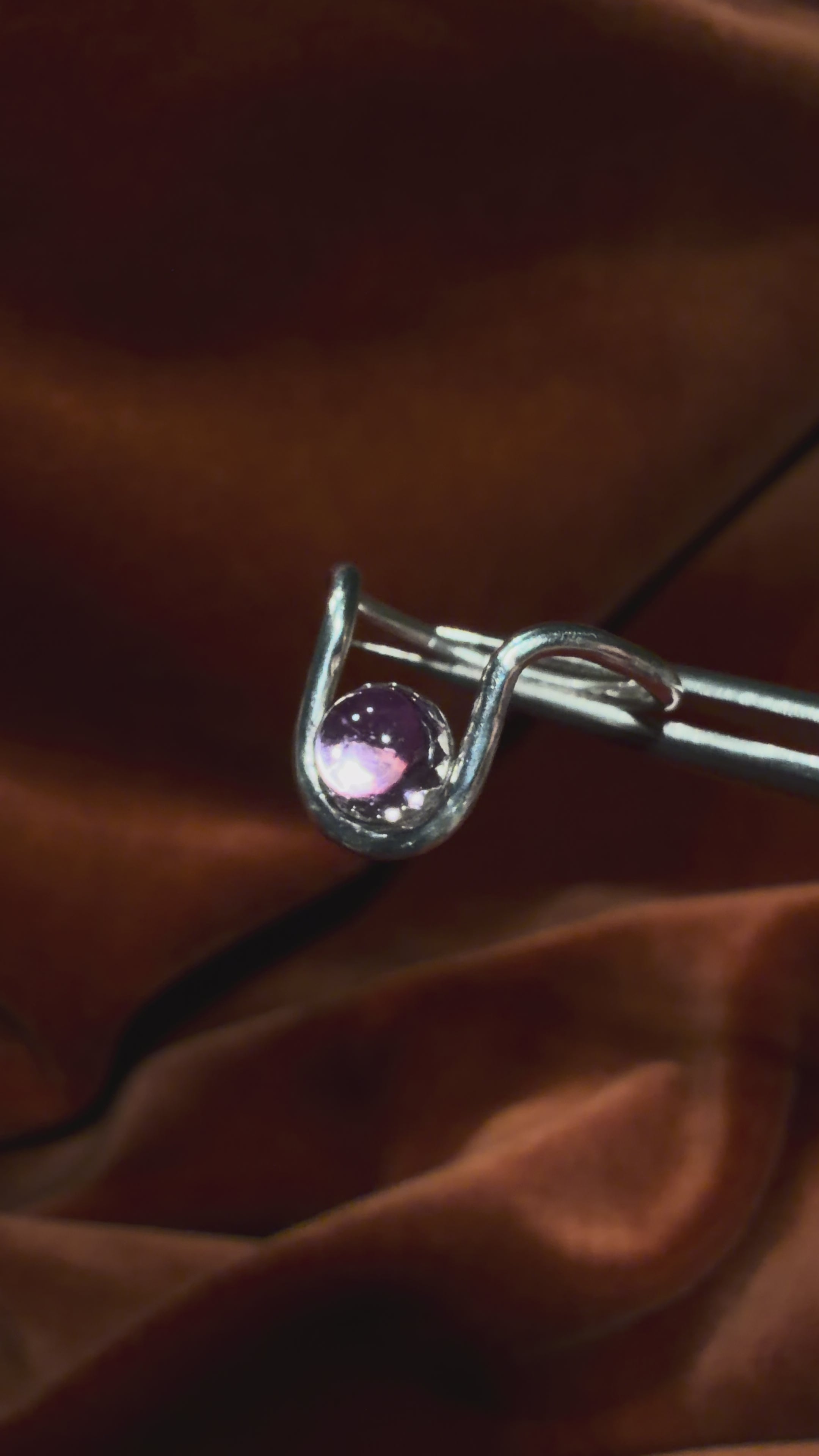 Close up video of a silver ring with bend in the ring where a serrated bezel holds a pink sapphire stone. Staged against an orange velvet background.