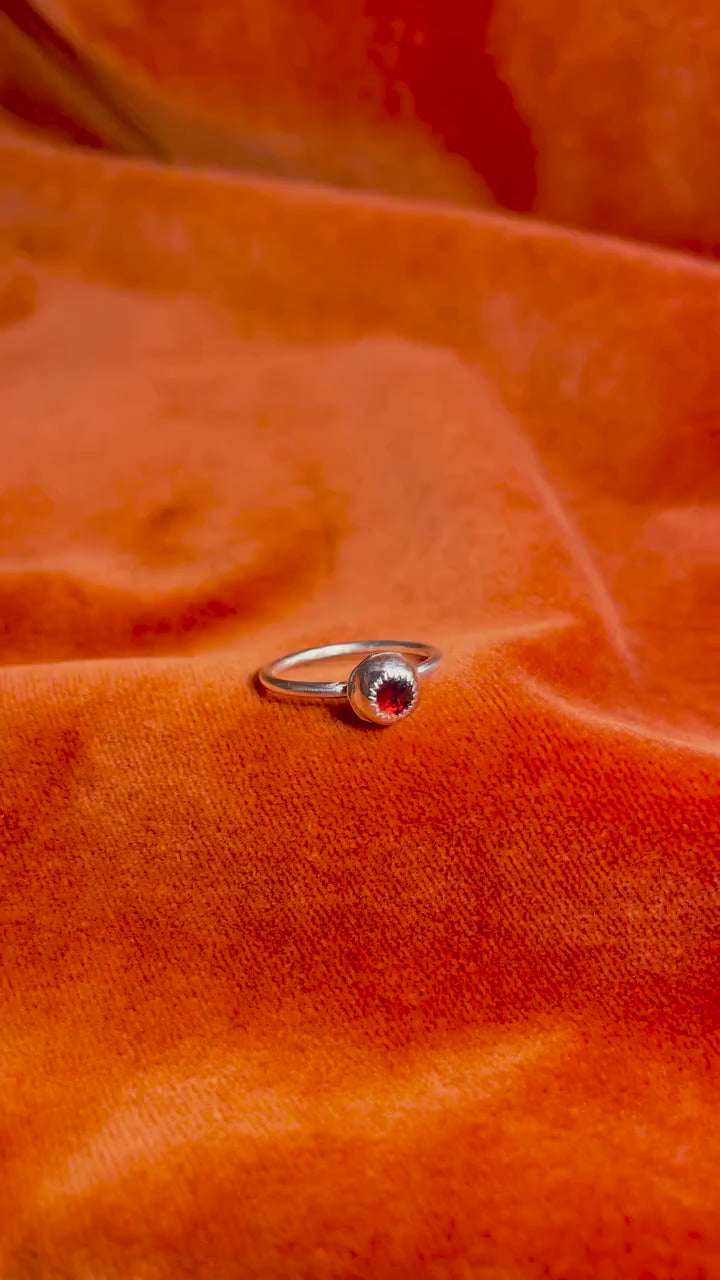A model's hand holds a silver ring with a serrated bezel enclosure and a rose cut garnet stone set inside sitting on a thin band. Ring is staged on an orange velvet background.