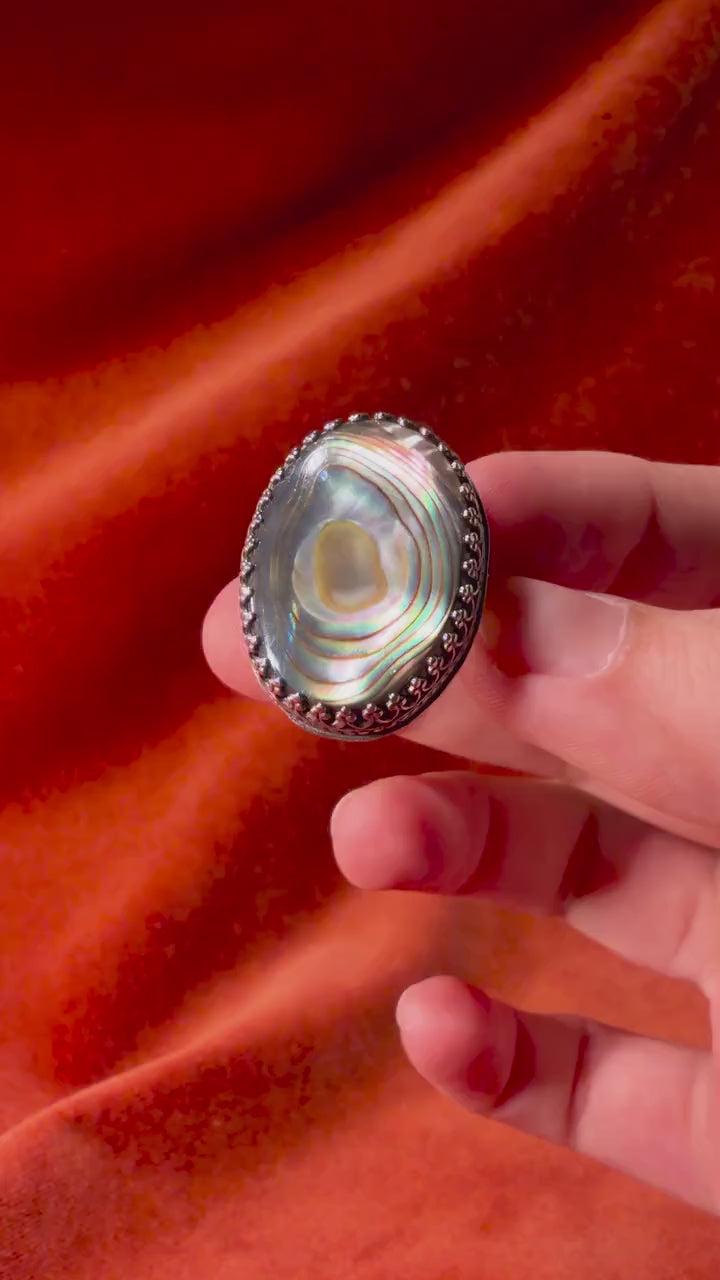 A model's hand moving a silver ring with a colorful abalone oval shaped stone set in an ornate bezel sitting on a medium sized band. Staged on an orange velvet background.