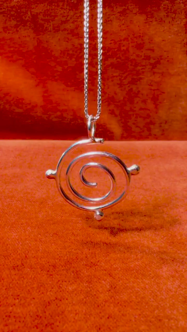 Silver spiral charm featuring small orbs. Pendant dangles on necklace over orange velvet background.