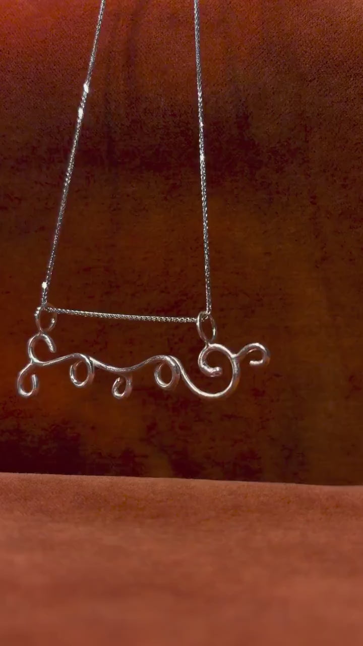 Video of a silver charm with wind swirls dangling from a silver chain. Staged against an orange velvet background.