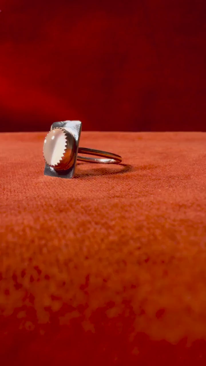 Video of a silver ring with a serrated bezel with a white moonstone set on a rectangular fixture attached to the band of the ring. Staged on an orange velvet background.