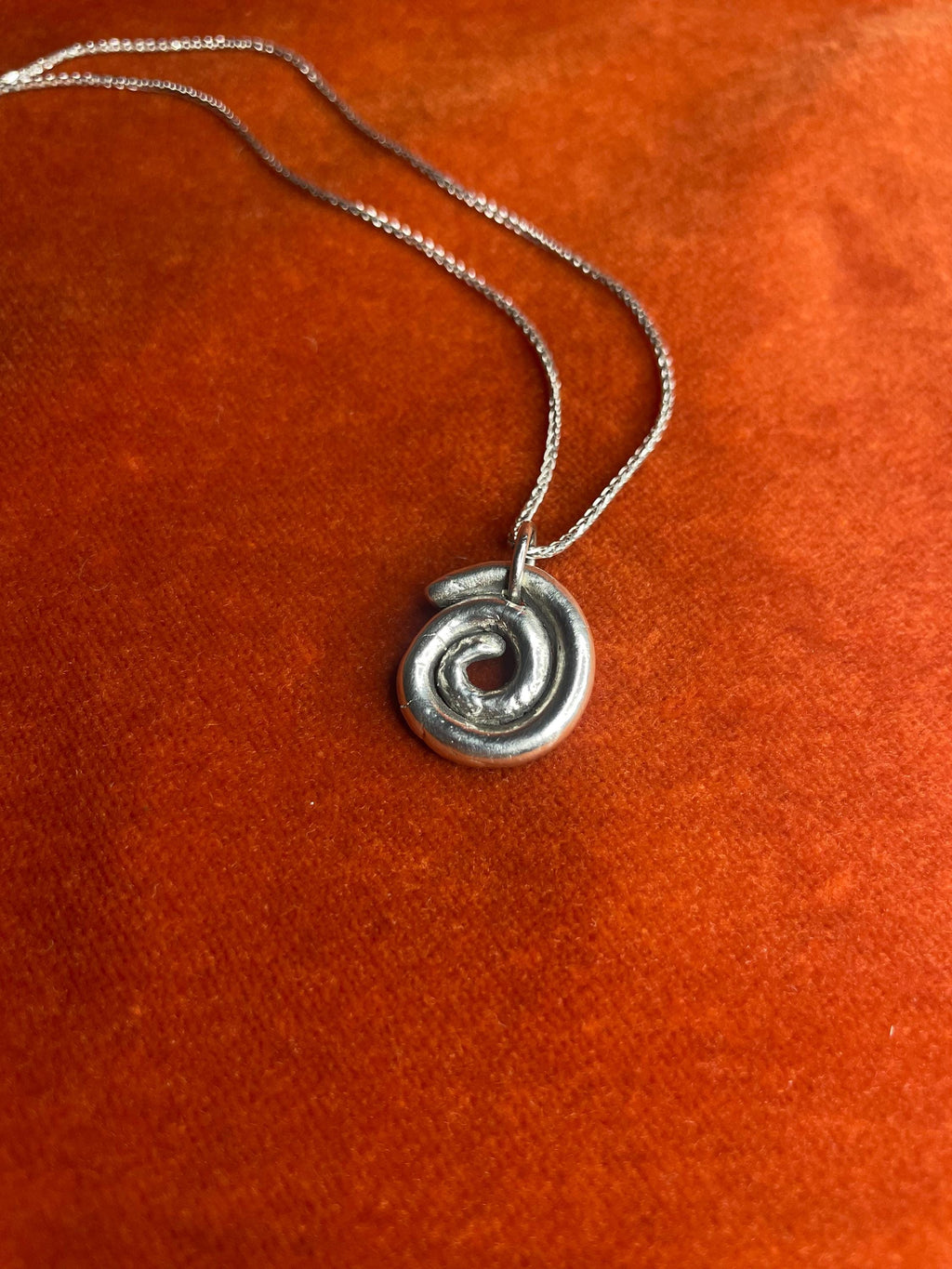 Silver spiral charm on a silver chain laying on an orange velvet background.