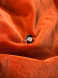 Image of a silver ring with a round rainbow white gemstone set on a square piece of silver over the band. Staged on an orange velvet background. 