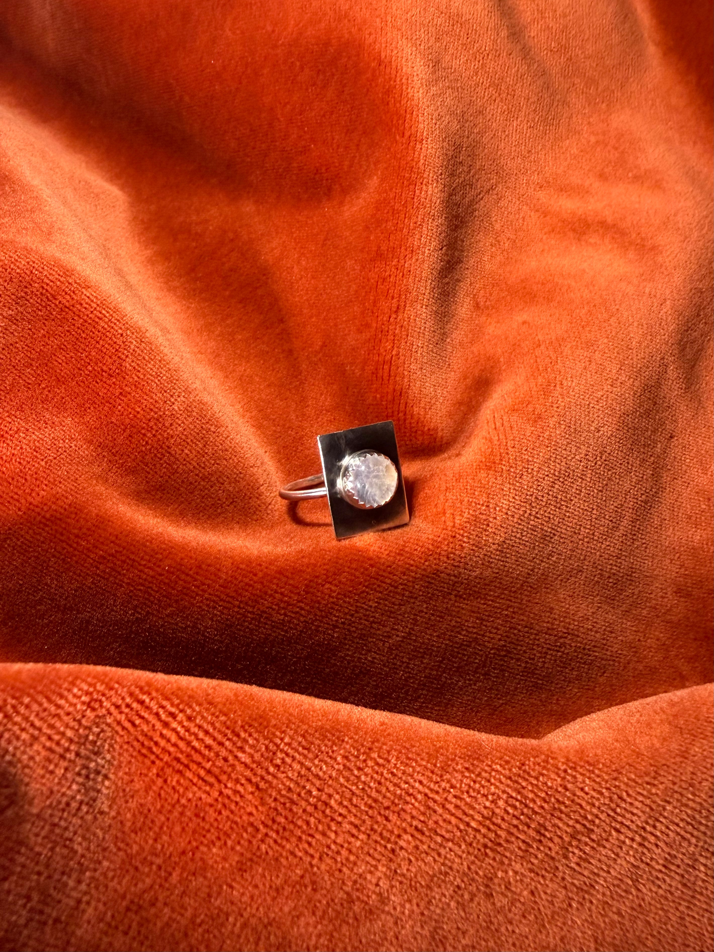 Image of a silver ring with a round rainbow white gemstone set on a square piece of silver over the band. Staged on an orange velvet background. 