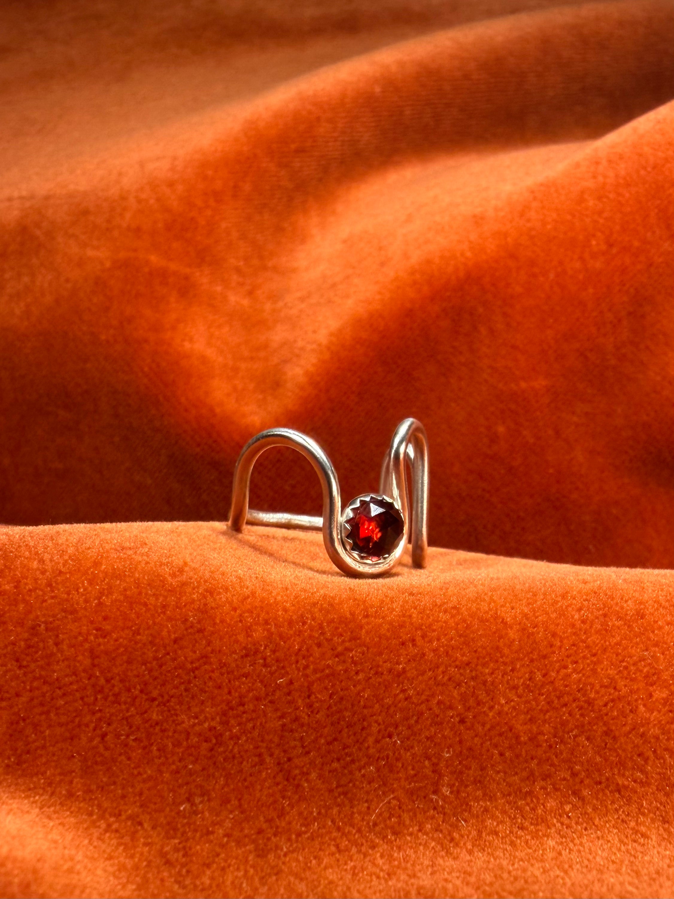 Close up image of silver ring with a asymmetrical band. A red rose cut garnet stone is set in a serrate bezel floating between the wavy band. 